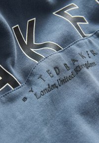 Modrá bavlněná tkanina s výrazným bílým a černým písmem. Text zní "Ted Baker" a "Londýn, Spojené království," s hladkou strukturou.