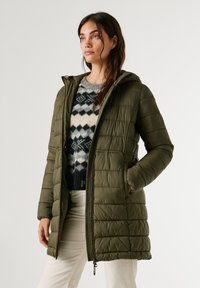 Oliņas zaļš pūkainais mētelis ar rāvējslēdzēja priekšpusi, kapuci un quiltotu tekstūru. Valkāts virs rakstainā džempera, kas ir taisnas formas un ar sānu kabatām.