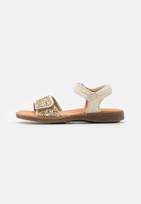 Sandales réglables avec un dessus en cuir beige, des accents en paillettes dorées et une semelle en caoutchouc marron. Elles disposent d'une sangle en velcro pour un maintien sécurisé.