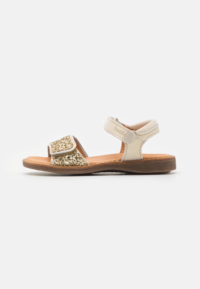 Sandales réglables avec un dessus en cuir beige, des accents en paillettes dorées et une semelle en caoutchouc marron. Elles disposent d'une sangle en velcro pour un maintien sécurisé.
