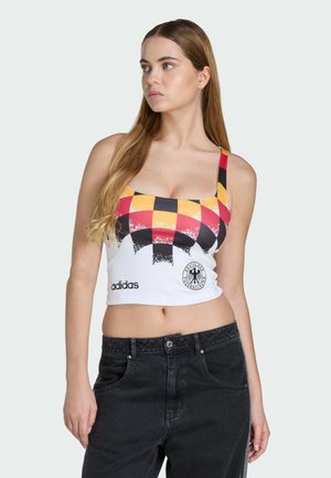 Młoda kobieta z długimi włosami, ubrana w bezrękawnik marki Adidas z czerwono-żółto-czarnym wzorem w kratkę oraz czarne dżinsy, patrząca w lewo.