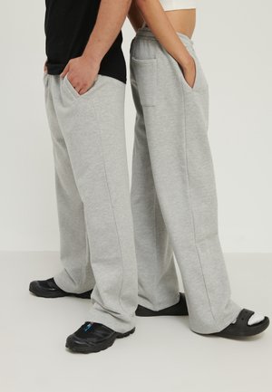 Deux personnes portant des pantalons de jogging larges gris clair, debout dos à dos, les mains dans les poches, l'une en baskets noires, l'autre en sandales noires avec des chaussettes blanches.