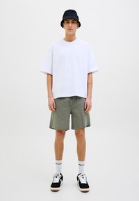 Camiseta blanca de gran tamaño, pantalones cortos verde oliva, sombrero de cubo negro, zapatillas negras y blancas, y calcetines blancos con diseño de logo.