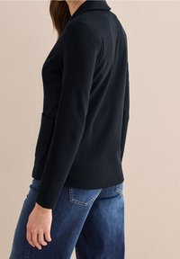 Černý pletený blazer s výstřihem do klopy, předními kapsami a přizpůsobeným střihem, v kombinaci s modrými džíny uvolněného střihu.