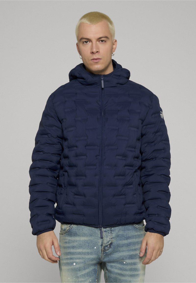 Junger Mann mit kurzem blondem Haar, der eine marineblaue gesteppte Kapuzenjacke und hellblaue ausgewaschene Jeans vor einem grauen Hintergrund trägt.