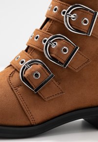 Brązowy zamszowy ankle boot z trzema metalowymi klamrami i akcentami w postaci pętelek, z czarną gumową podeszwą i teksturowaną powierzchnią.