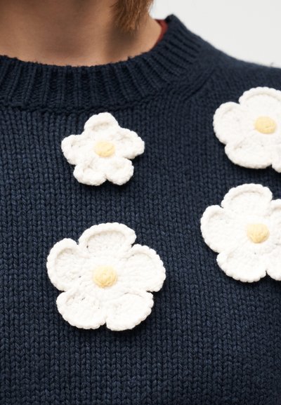 Marineblauer Strickpullover mit fünf weißen Häkelblumen mit gelben Zentren, die in Größe und Textur variieren und gleichmäßig über die Vorderseite verteilt sind.
