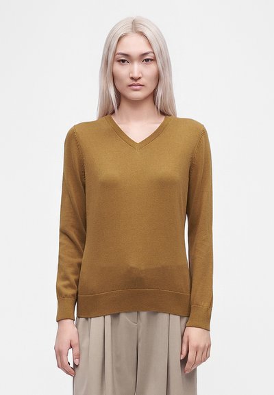 Jeune femme aux longs cheveux raides platine portant un pull à col en V jaune moutarde et un pantalon large beige clair, debout devant un fond blanc.