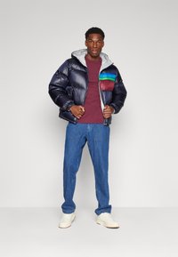 Chaqueta tipo puff en azul marino oscuro con capucha gris, con una colorida franja horizontal en el pecho, combinada con pantalones de mezclilla azul y zapatillas blancas.