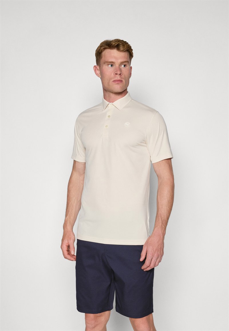 Hellbeige Poloshirt mit einem kurzen Kragen, drei Knöpfen und dezentem Logo. Kombiniert mit marineblauen Shorts, die einen glatten Stoff haben.