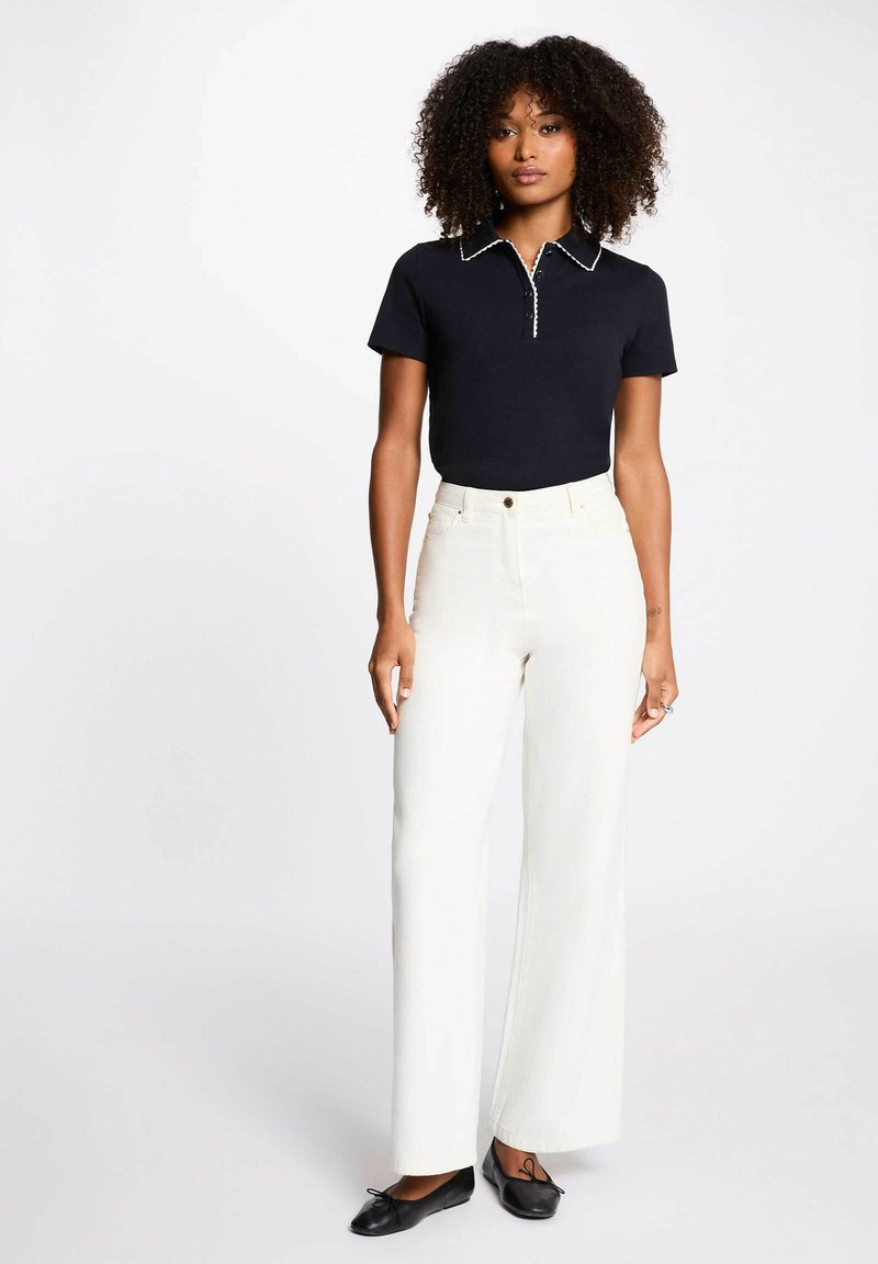 Donna con capelli ricci che indossa una polo nera a maniche corte, pantaloni bianchi a vita alta e gamba larga, e ballerine nere, in piedi su sfondo bianco.