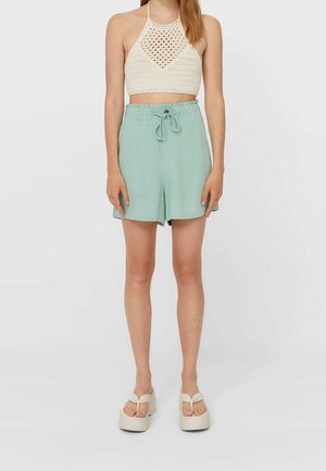 Woman wearing a white crocheted halter crop top, light green high-waist shorts with a drawstring, and white platform sandals.