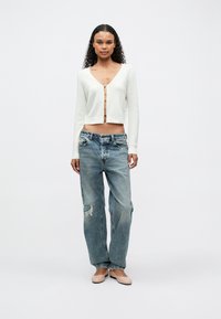 Mujer con cabello largo y rizado, vistiendo un cárdigan corto blanco con botones de letras, jeans rotos y holgados, y bailarinas beige, de pie sobre un fondo blanco.