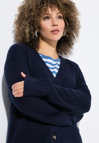 Gilet tricot navy à manches longues et décolleté en V profond, avec une texture lisse et une fermeture par un bouton unique. Chemise rayée visible en dessous.