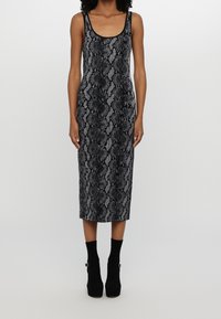 Femme portant une robe midi sans manches imprimée serpent noire et grise avec des bottines noires sur un fond clair uni.