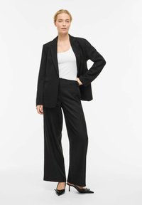 Črni krojeni blazer in široke hlače, kombinirani z belo majico brez rokavov. Črni čevlji s koničastim prstom in pentljo. Gladek, strukturiran material.