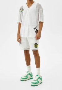 Valkoinen verkkomallinen lyhythihainen paita napituksella, yhdistettynä sopiviin shortseihin, joissa on värikkäitä korjauksia. Ulkoasun täydentävät vihreä- ja valkopohjaiset kuviolliset tennarit.