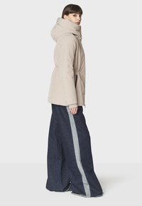 Chaqueta aislante beige con capucha, que presenta una cintura ajustada, bolsillos laterales y se combina con pantalones de mezclilla de pierna ancha con franjas laterales grises.