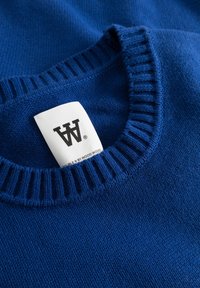 Pull en maille bleu avec un col côtelé et une étiquette portant la mention "DOUBLE A BY WOOD WOOD." La texture est douce avec une finition lisse.