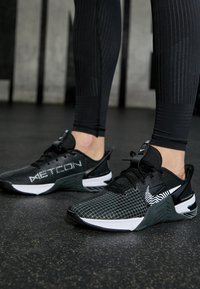Sorte sportsko med mesh-overdel, prydet med et hvidt "METCON" logo, teksturerede accenter og et særpræg design med striber på siden.