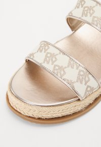 Sandaler med to brede kremfargede stoffstropper som har et subtilt gullmonogrammønster, metallisk rosagull fotbrett og sål innpakket i jute.