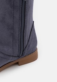 Zbliżenie ciemnoniebieskiego zamszowego buta z widocznym bocznym zamkiem błyskawicznym i brązowym niskim obcasem na białym tle.