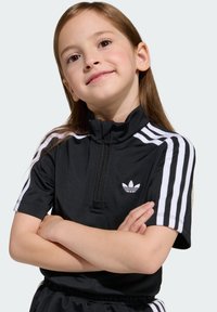 Čierny športový top na zips s krátkymi rukávmi, bielymi troma pruhmi a malým bielym logom Adidas na ľavej strane hrudníka. Mäkká textúra látky.