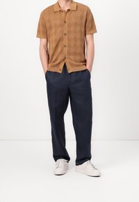 Kortsleeve, rutmönstrad skjorta i brunt med krage, tillsammans med marinblå byxor. Vita sneakers fulländar outfiten.