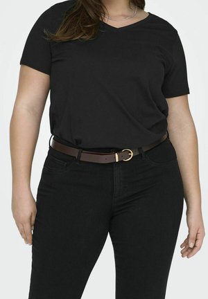Frau trägt ein schwarzes T-Shirt mit V-Ausschnitt, schwarze Jeans und einen braunen Ledergürtel mit goldener Schnalle vor einfarbigem Hintergrund.