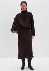 Sweatshirt zippé marron avec col haut, associé à une jupe marron. Le modèle tient un sac marron texturé. Des bottes noires jusqu'aux genoux viennent compléter le look.