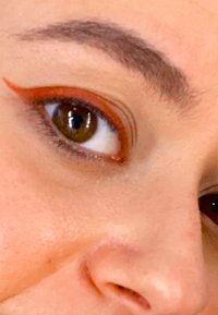 Gros plan sur un œil avec un iris brun foncé. Présente un eyeliner orange proéminent sur la paupière supérieure, sur un teint de peau clair et un sourcil.