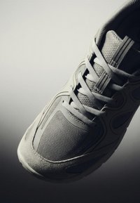 Baskets de sport gris clair avec des panneaux en mesh et en daim, sécurisées avec des lacets plats assortis, présentées sur un fond gris dégradé.