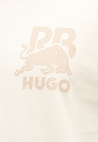 Λογότυπο με άλκιμο ταύρο, με τα γράμματα "RB" από πάνω και τη λέξη "HUGO" από κάτω, σε φόντο ελαφρώς χρωματισμένου υφάσματος.