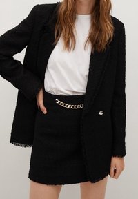 Blazer en tweed noir avec bords effilochés, associé à une jupe assortie ornée d'une ceinture chaîne dorée. Texture douce et design structuré.