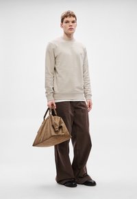 Μπεζ φούτερ με ριμπ μανσέτες, καφέ παντελόνι με φαρδιά μπατζάκια και καφέ δερμάτινη τσάντα. Το μοντέλο στέκεται μπροστά σε ανοιχτό γκρι φόντο.