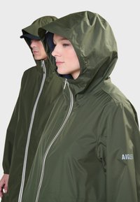 Veste imperméable verte avec capuche, dotée d'une fermeture éclair sur le devant, de poignets élastiques et d'un logo sur la manche. Tissu lisse et léger.