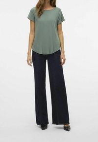 Top verde de manga corta con cuello redondo y un diseño fluido, combinado con jeans de pierna ancha oscuros y zapatos negros de punta.