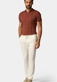 Rustkleurige korte mouwen polo met verticale ribbels, gecombineerd met crèmekleurige broek met een trekkoord. Model draagt beige sneakers.