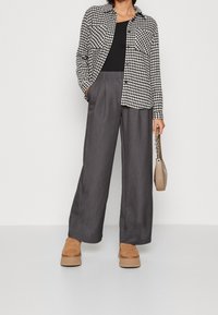 Giacca camicia a scacchi bianco e nero, top aderente nero, pantaloni ampi grigi, scarpe con plateau marroni, in mano una borsa beige.