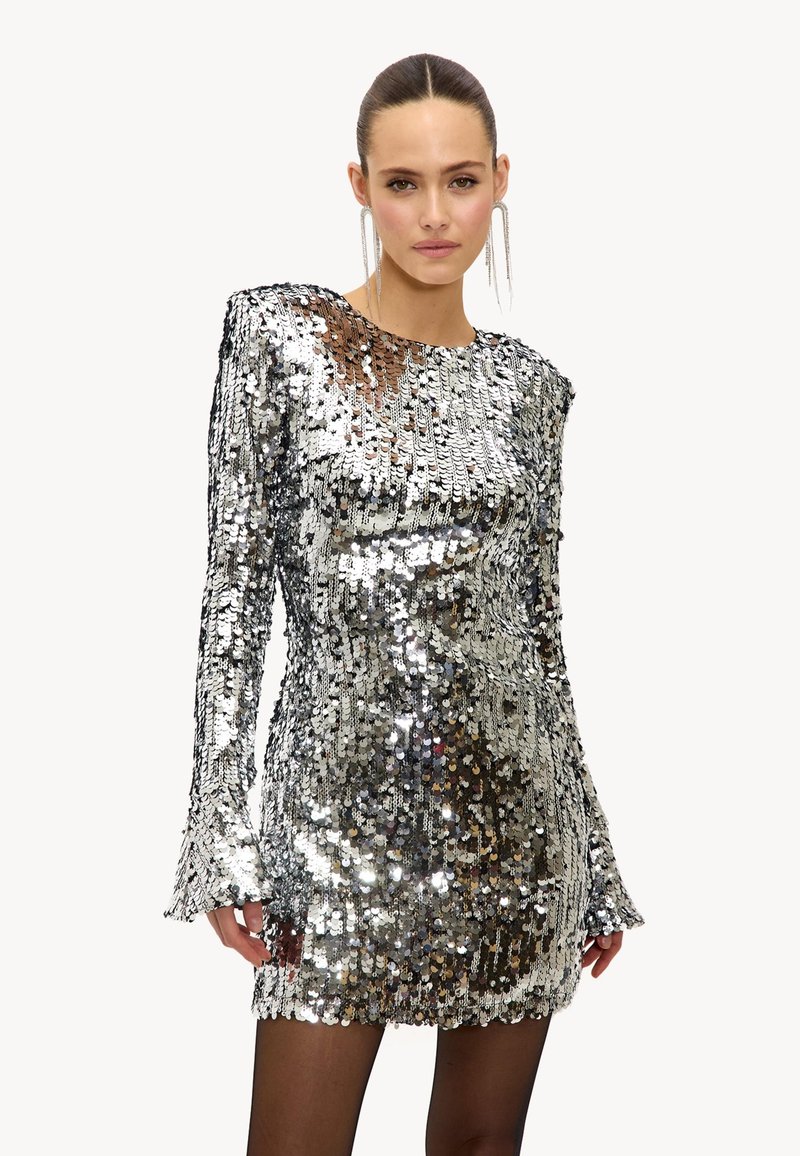 Femme portant une mini-robe argentée brillante à sequins avec des manches longues et des boucles d'oreilles remarquables, posant devant un fond blanc uni.