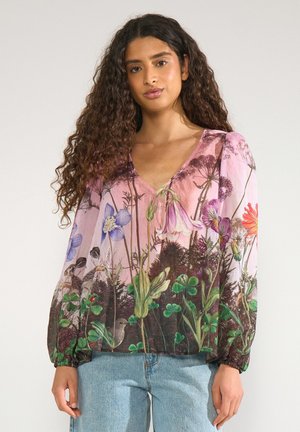 Kvinde med langt krøllet hår har en lyserød bluse med farverigt blomsterprint og lyseblå jeans på og står mod en ensfarvet baggrund.