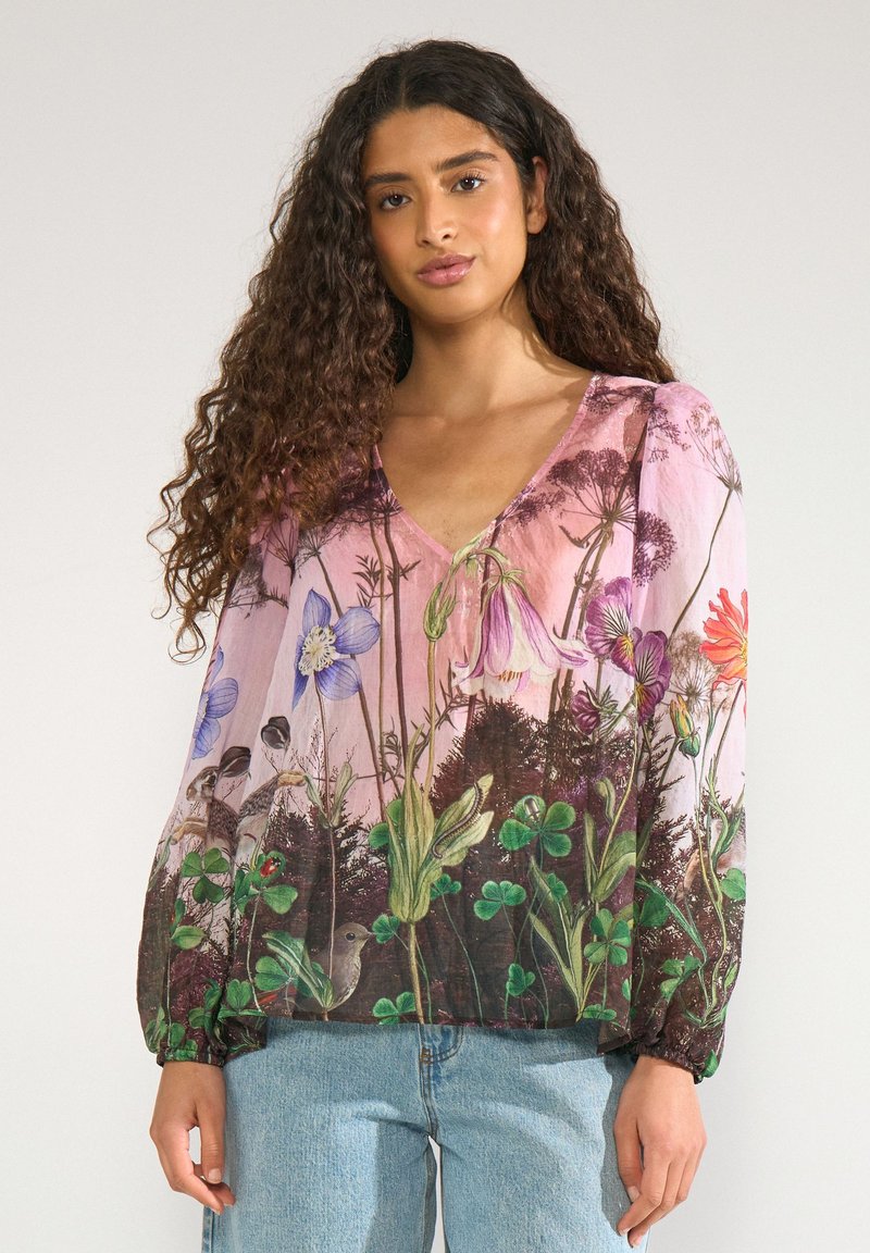 Femme aux longs cheveux bouclés portant un chemisier rose à imprimé fleuri coloré et un jean bleu clair, debout sur un fond uni.