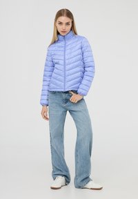 Giacca trapuntata di un lieve viola con colletto alto, caratterizzata da una chiusura con zip e un motivo a zigzag. Abbinata a jeans blu larghi e sneakers bianche.