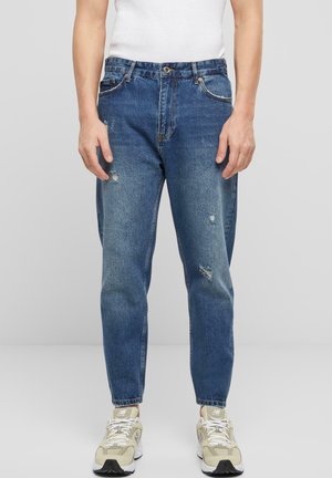 Mann trägt blaue, verwaschene Jeans, weißes Tanktop und beige Sneaker, steht vor einem schlichten hellgrauen Hintergrund.