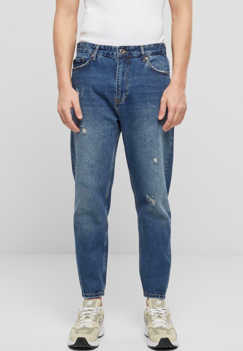 Homme portant un jean bleu usé, un débardeur blanc et des baskets beiges, debout devant un fond gris clair uni.