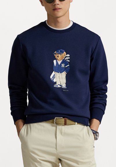 Sweatshirt bleu marine à col rond présentant un graphisme d'ours en tenue de golfeur, avec des détails brodés et une étiquette logo au bas.