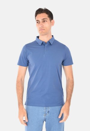 Uomo in piedi, rivolto verso l'obiettivo, che indossa una polo blu a maniche corte e jeans azzurri chiari su uno sfondo chiaro e uniforme.