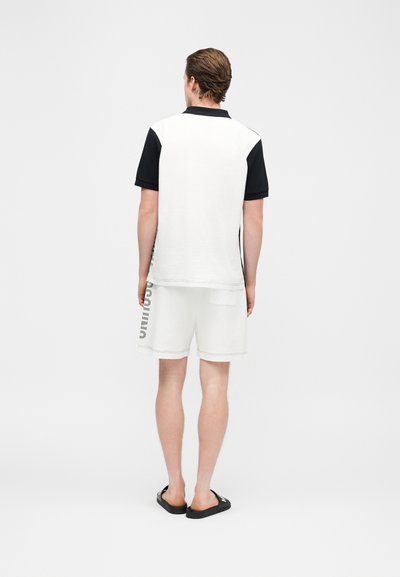 Homme debout, vu de dos, portant une chemise à manches courtes blanche et noire, un short blanc avec un motif sur le côté, et des sandales noires à enfiler.