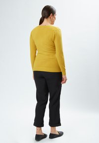 Senfgelbes, eng anliegendes Sweatshirt mit langen Ärmeln, geripptem Saum, kombiniert mit schwarzen hochgekrempelten Hosen und schwarzen flachen Schuhen.