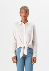 Lys rosa skjorte med knapper og frontknute-detalj, lange ermer, parret med blå tettsittende bukser. Materialet ser mykt og teksturert ut.